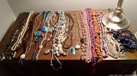 Photo of assorted necklaces laid out on wooden surface seen from above, showing multiple bead styles and colors including blue, white, pink, and brown. Heno watch pendant visible.