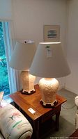 Pair of two tall Asian style ceramic table lamps on a wooden table, with cream fabric shades and carved floral details on lamp bases.