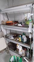 Full side view of the metal plant stand filled with miscellaneous gardening and household items. Shows two shelves with long white grow light fixtures above each shelf, silver metal frame with A-frame side supports.