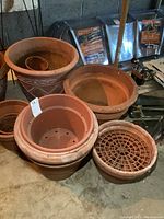 Grouped view of multiple clay and resin planters in various sizes and styles on a concrete floor in garage setting.