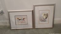 Photo showing two framed signed prints side by side on the floor: one larger featuring stylized birds, the other smaller featuring a group of birds.