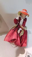 Front view of Royal Doulton figurine showing detailed face, orange-red hat with blue ribbon, red dress, and green scarf.