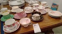 Overview showing arranged collection of twelve assorted vintage teacups and saucers in floral and decorative patterns on a wood table.