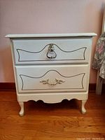 Front view of white wood bedside table showing two drawers, decorative handles, and scalloped base with curved legs.