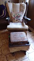 Wood rocking chair with beige fabric seat and back cushion, floral pillow on the seat, and visible scratches on the wooden arms. Matching ottoman with patterned cushion in front.