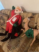 Santa Claus figurine in red suit holding a black toy bag, sitting on patterned couch with geometric pillows.