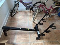 Photo showing the full bike rack lying on floor next to bicycles. Black metal with mounting hardware and support arms visible.