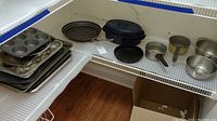 Photo of multiple aluminum baking pans, muffin tins, perforated broiler tray, Wilton cookie sheet, small saucepans and mixing bowls on white wire shelving.