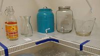 Shelf view showing 2 glass beverage bottles with floral design, a blue metal dispenser, clear glass dispenser with metal lid, and clear abstract glass bowl to the right.