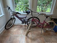 Two children's bicycles in a sunroom with tile floor near windows, showing overall front and side views of both bikes.