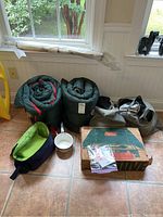 Full lot arranged in a room near windows showing green rolled sleeping bags, cooler bag, enamel pots, stove box, umbrella, and bags containing inflatable beds.