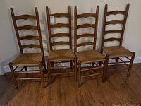Four solid wood ladder back chairs with rush woven seats and stretcher supports shown front-facing in a room with wood flooring.