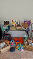 Wide view of table with children's books, puzzles, toys, plastic bins, and wooden crates along with an Avengers-themed suitcase on floor.