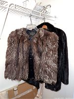 Two mink fur jackets hanging on hangers, one brown multi-toned and one black, visible hanging on a white wire shelf.