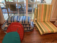 Photo showing multiple outdoor and indoor chair cushions stacked near a sliding glass door, including green striped, blue striped, red and teal cushions.