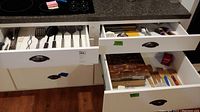 Wide view of three kitchen drawers showing assortment of kitchen utensils, knives, spoons, cutting boards, and spice racks