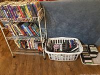 Photo showing over 40 VHS tapes on a white metal rack, plus a plastic laundry basket containing DVDs and a box of cassettes nearby. Tapes include various Disney titles and classic movies.