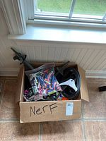 Box filled with four Nerf shooters, visible darts bag and two black targets inside near a window on tile floor.