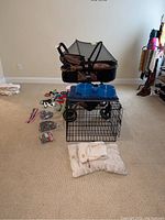Wide view showing the black folding crate, blue plastic dog bowls on top, various dog toys and harnesses laid out, and brown/black pet stroller with canopy in background.