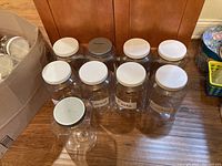 Several medium-sized clear plastic storage jars with white lids shown on a wooden surface. Some jars labeled for kitchen storage.