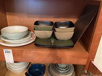 Multiple stacked plates and bowls including floral patterned round plates, solid colored square bowls, and rectangular platter in green.