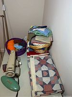 Photo showing stacked folded hand-stitched quilt, basket of scrap fabrics, skeins of yarn in a basket, and a vintage sewing machine with green iron accessory beside them.
