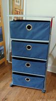 Front view of metal rolling shelf with four blue canvas drawers showing circular metal grommet handles and metal frame with stains.