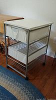 Side angle showing steel frame, white top, two cloth drawers and two metal wire basket drawers on caster wheels