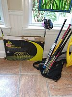 Photo showing five aluminum softball bats held upright in a black carrying bag, with a box of soccer passing arches next to them on a tiled floor by windows.