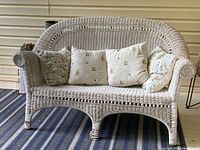 Front view of white wicker loveseat with four pillows on a porch showing overall shape and cushion design.
