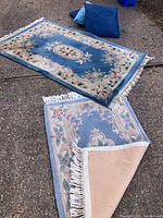 Photo showing two floral patterned blue area rugs laid on the ground with two blue throw pillows behind them. Rugs have fringed edges and visible stains.
