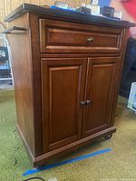 Front and angled view of wooden cabinet with black stone top, showing drawer, door fronts, metal knobs and wheels.