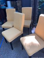 Four fabric covered straight back chairs with light yellow upholstery and dark wooden legs, photographed outdoors on pavement near garage.
