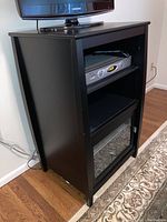 Front-right view of black wooden entertainment chest showing glass door, adjustable shelf with a digital cable box inside and TV on top.