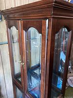 Front upper section of the curio cabinet showing wooden frame, glass front and sides, brass handles, and decorative arches.