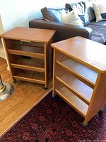 Two matching wooden end tables with three shelving tiers, on caster wheels, placed on a floor near a couch and carpet.