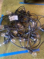 Coiled outdoor string lights showing multiple clear bulbs and black cable wiring on green carpet with blue tape marks.