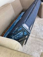 Sleeper sofa partially unfolded showing blue patterned mattress and metal fold-out bed frame inside beige cloth sofa.