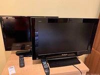 Two black 26 inch televisions side by side on a wooden surface with their remotes placed in front.
