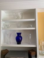 Shelving unit displaying seven glass items: one blue opaque vase centered on middle shelf, two tall clear vases on sides, and three small clear glass bowls on upper shelf.