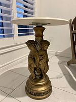 Side view of bronze pedestal base with two cherub figures holding an urn, detailed drapery and wings, supporting a round marble top.