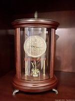 Front view of the Bombay Co. anniversary clock showing clock face with Roman numerals under a glass dome and wood finish.