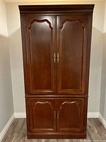 Front view of the dark brown wooden armoire cabinet with closed double doors, showing carved panel design and brass handles