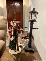 Photo showing the pair of ice skating figurines and the black lamp post on a wooden surface indoors.