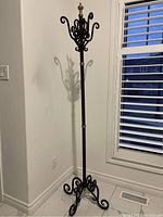 Full view of tall metal hall coat rack with curled hook design and brass top finial, standing on tiled floor.