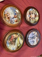 Four round collector plates with detailed painted scenes, each in a wooden frame, laid on red fabric surface