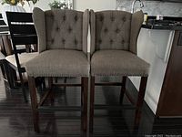 Two grey fabric-upholstered counter height chairs with button-tufted backs and dark wooden legs