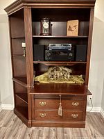 Full frontal view of the wooden display cabinet showing three display shelves and storage drawers below, placed against a wall, with various decorative items for scale reference.