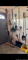 Photo showing various garden tools hanging on wall, including rakes, shovels, broom, and hand tools.