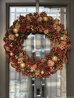 Front view of a decorative round wreath made with pinecones, gold glitter pinecones, gold balls, berries, and green leaves hanging on a glass door.